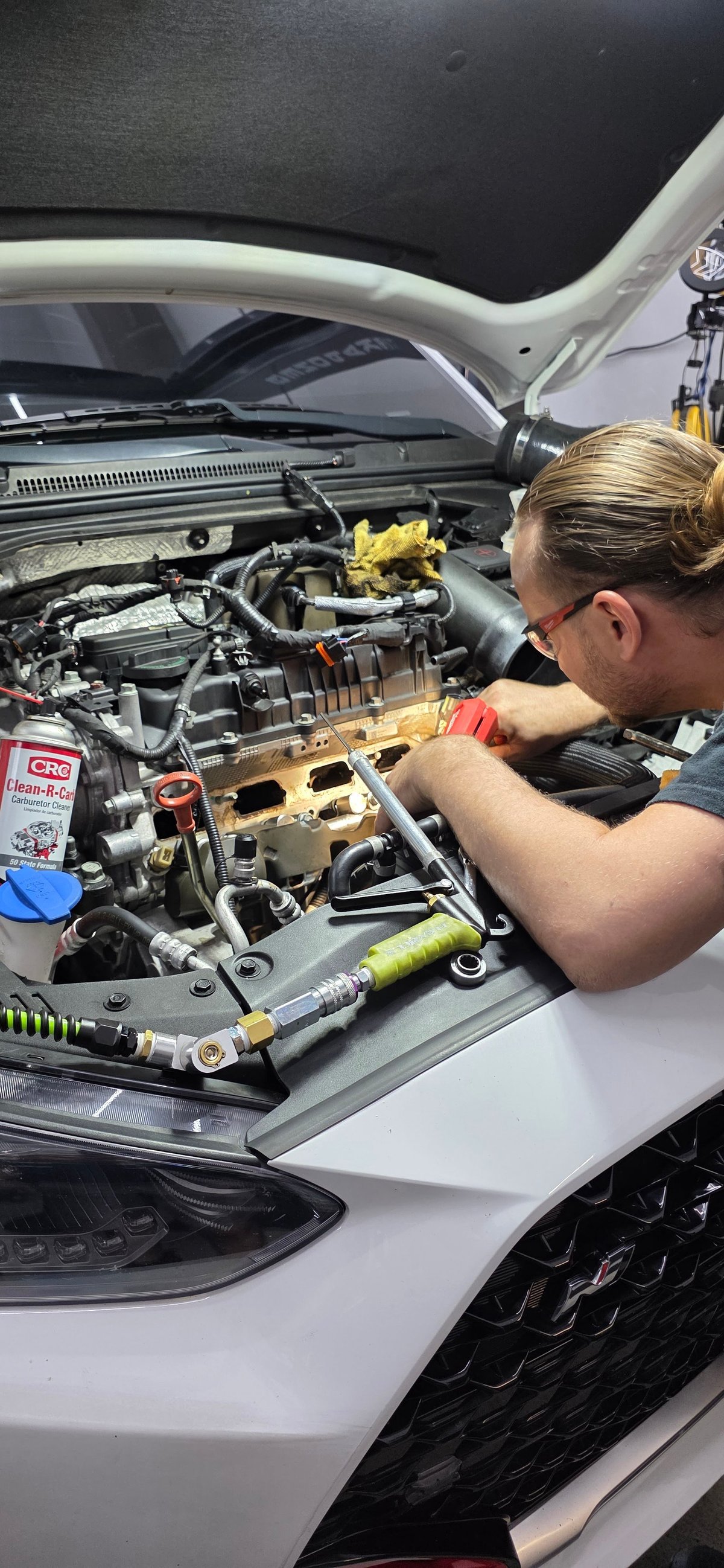 Mechanic working on white car engine compartment, leaning over open hood to inspect or repair engine components