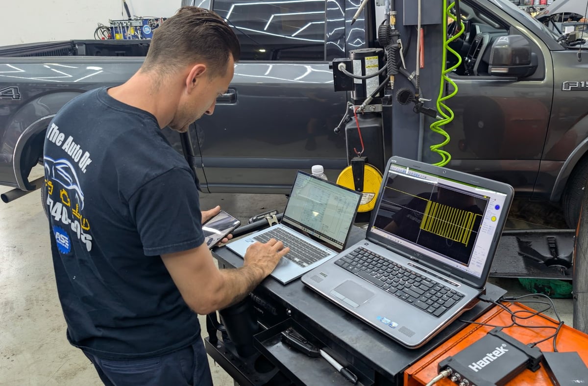 Technician working on laptops near open vehicle door with diagnostic equipment in outdoor garage setting